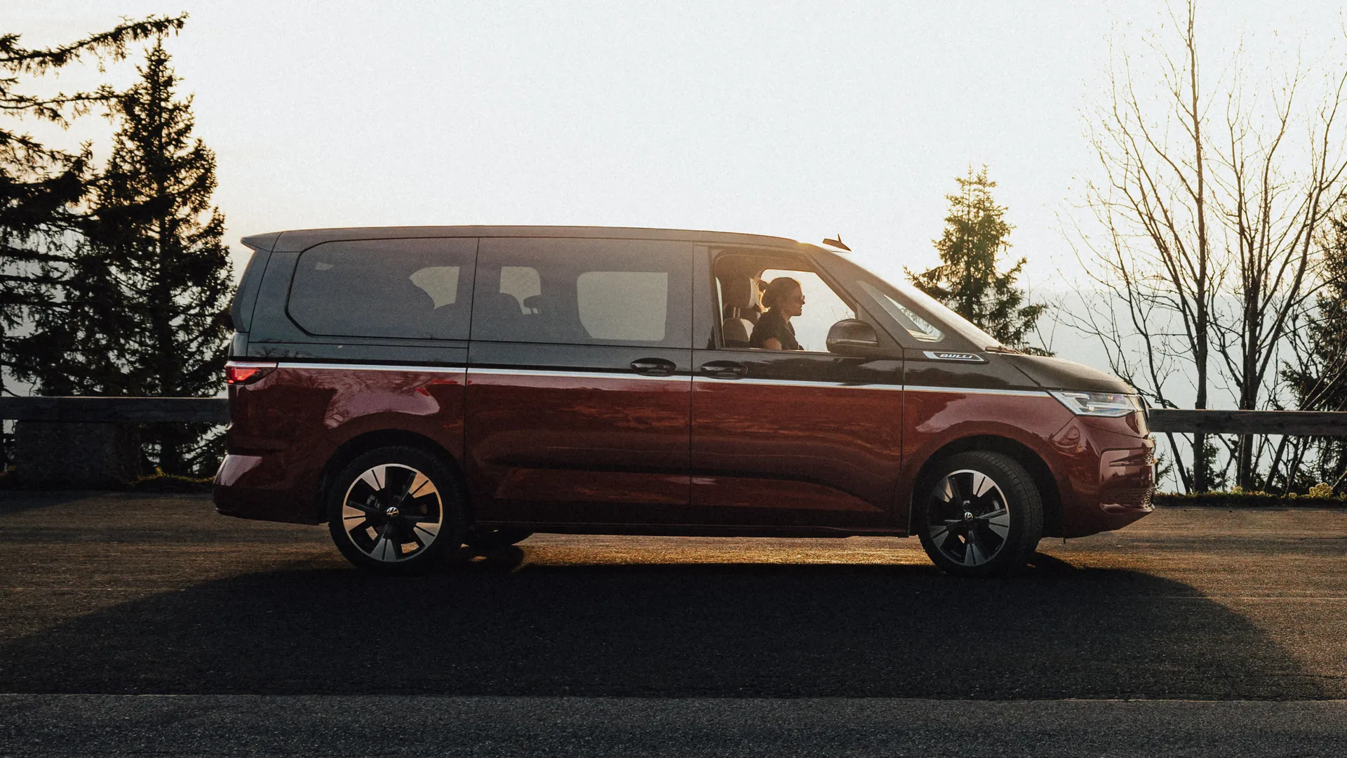 Vista lateral de Volkswagen T7 Multivan bicolor rojo-negro. Vista de lateral frente al atardecer